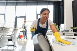 Smiling,Bi racial,Woman,Cleaning,Office,Desk,With,Rag,Near,Colleague